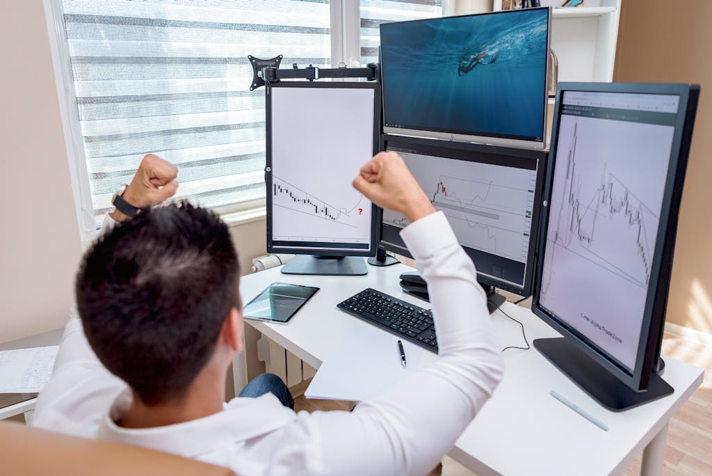 A man celebrates in front of multiple computer monitors displaying stock market graphs, indicating trading success.